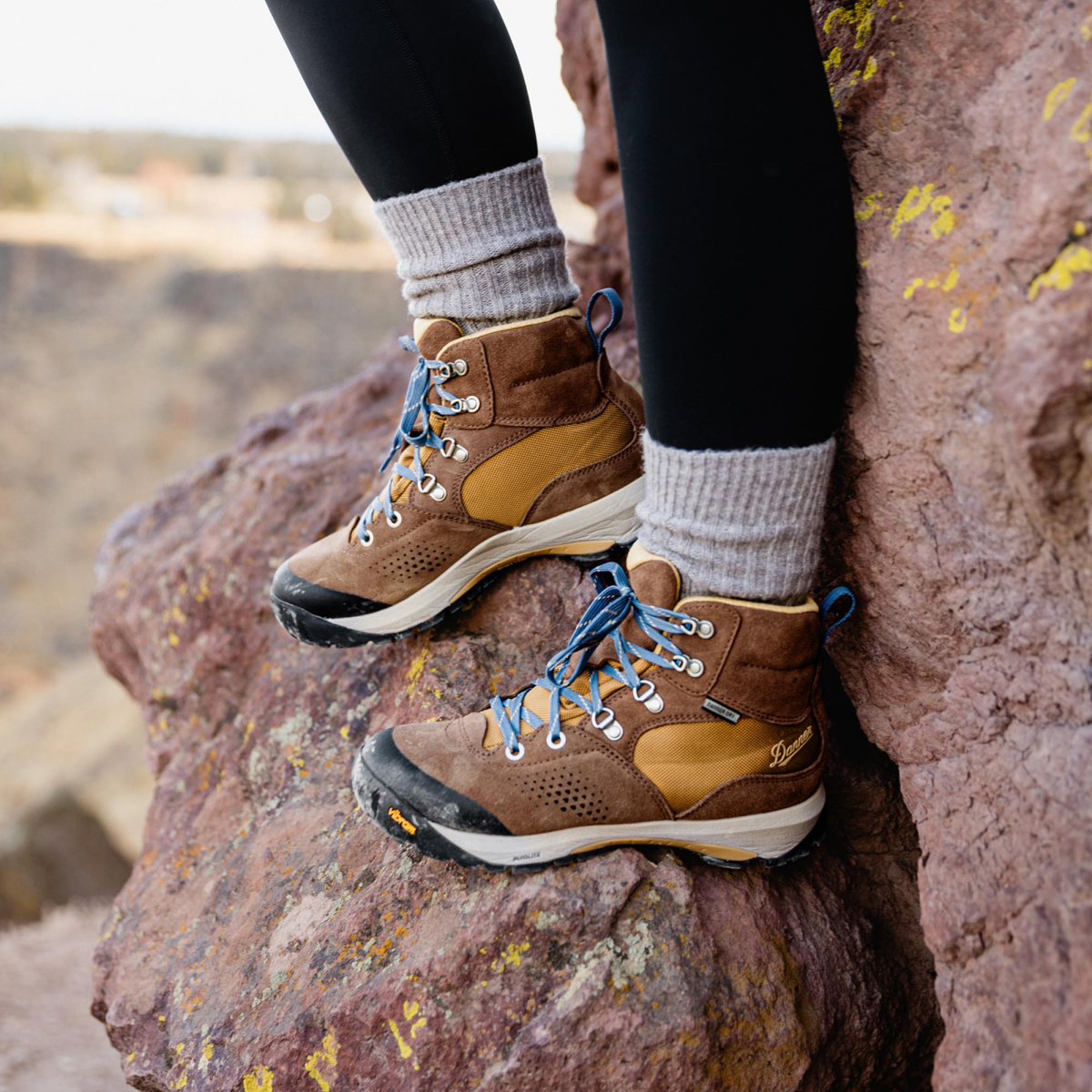 A woman pauses on a rock while hiking wearing black leggings, gray socks, and brown leather boots with yellow textile inlays and light blue laces.