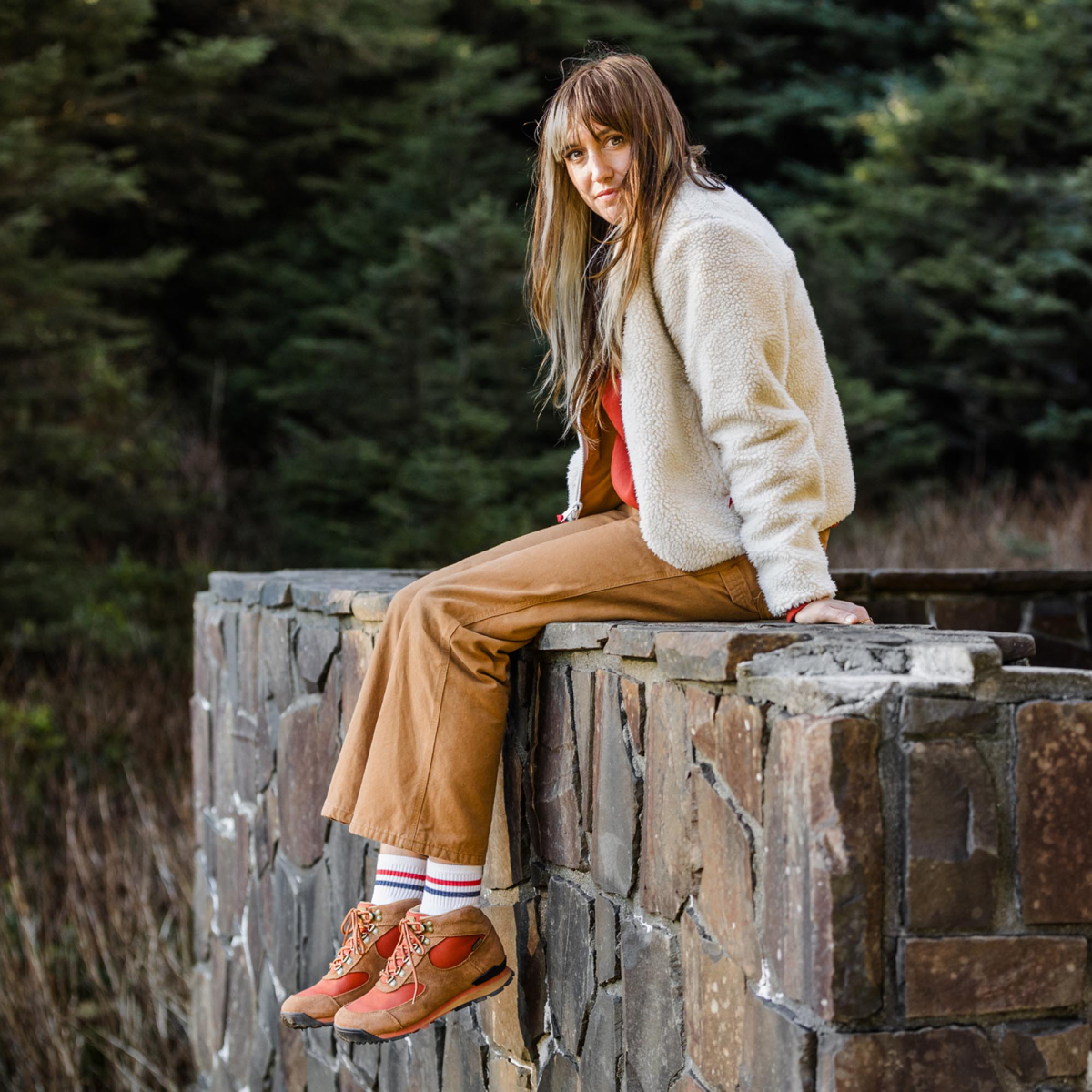 A woman sits on a rock wall, her legs dangling off the edge, wearing a white fleece top, tan pants, and brown leather boots with red textile inlays.